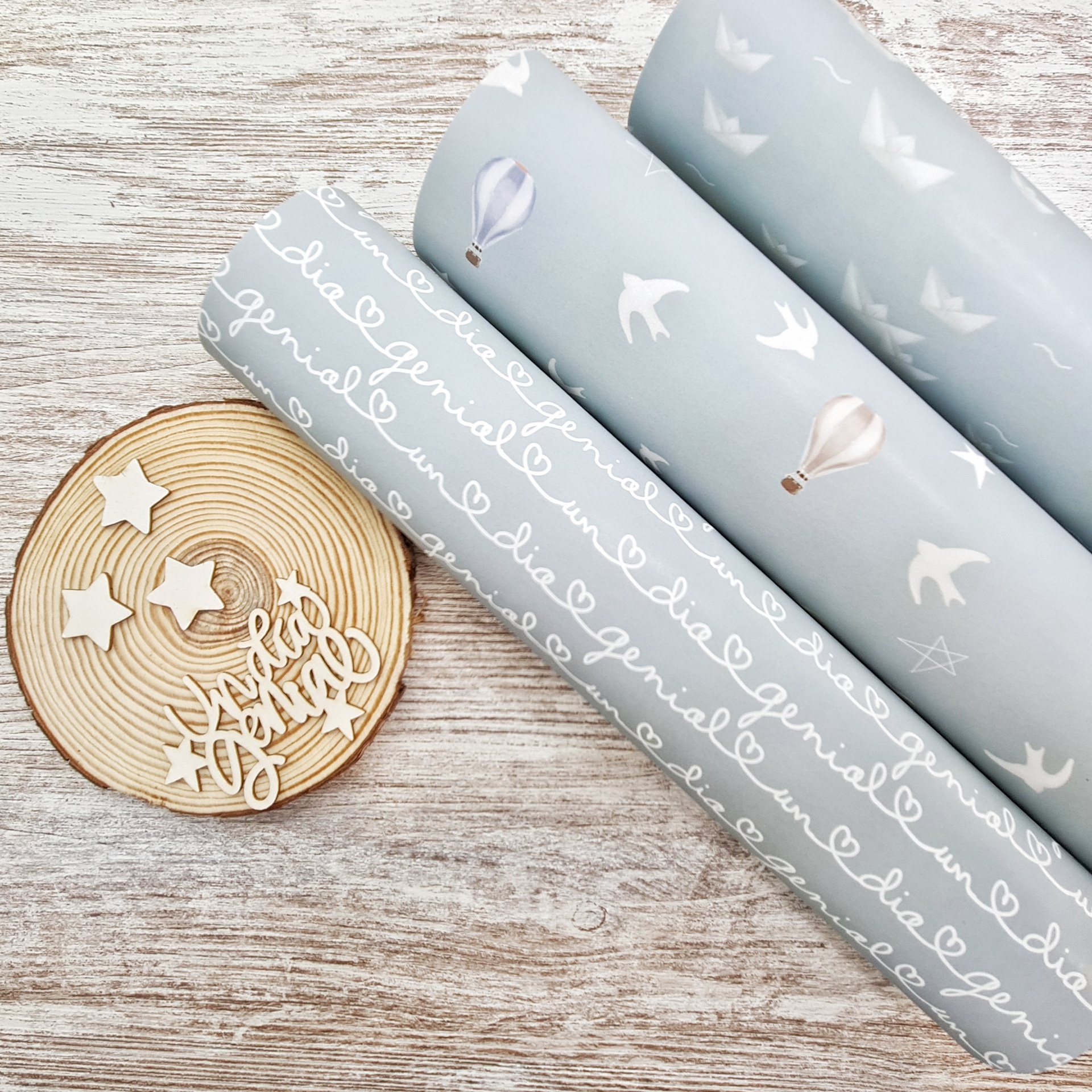 Três rolos de tecido azul claro com desenhos de pássaros, balões de ar quente e texto, e base decorativa em madeira com a frase Feito com Carinho.