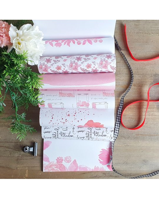 Rolos de papel decorativo com estampas florais em rosa e branco, fitas e flores sobre mesa de madeira.