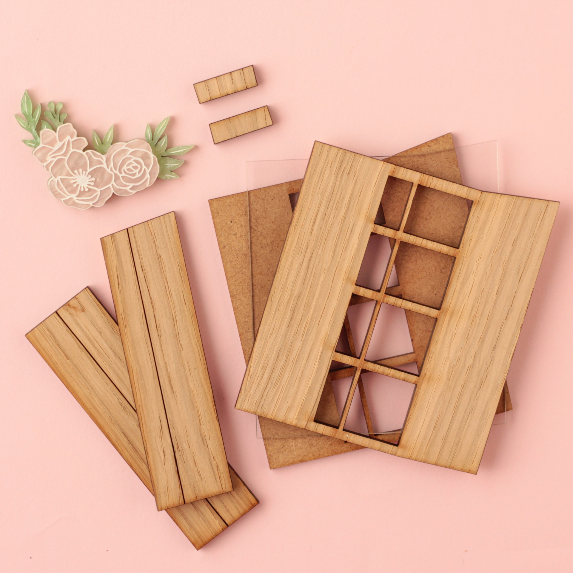 Peças de madeira clara desmontadas com aplicação decorativa de flores brancas e folhas verdes sobre fundo rosa