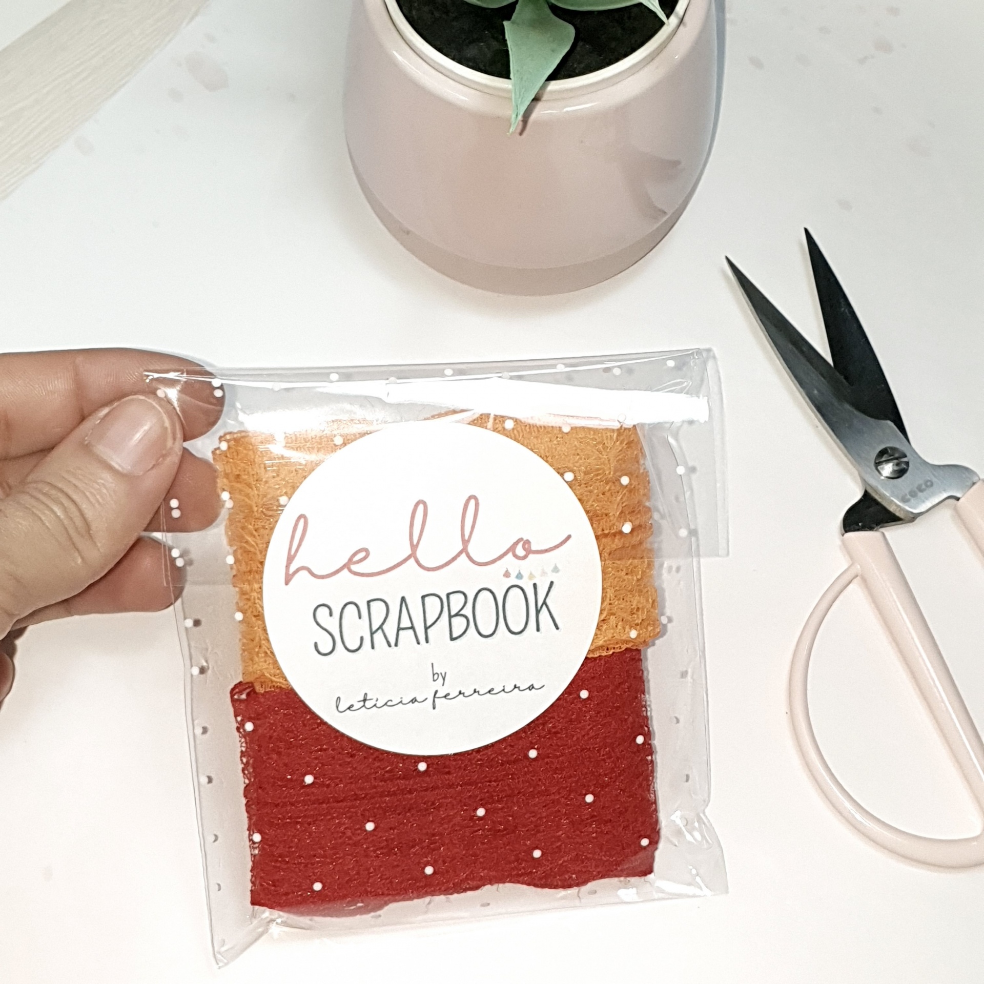 Saco transparente com tecidos castanho claro e vermelho com bolinhas, tesoura rosa e vaso bege claro