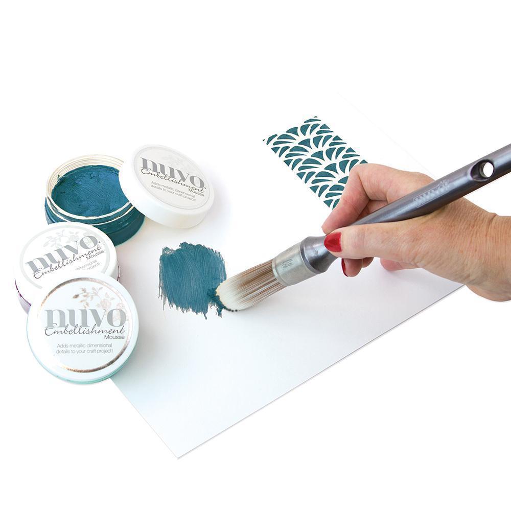Mãos aplicando tinta azul esverdeada da marca NUVO Embellishments com pincel grande em papel branco ao lado de padrão azul.