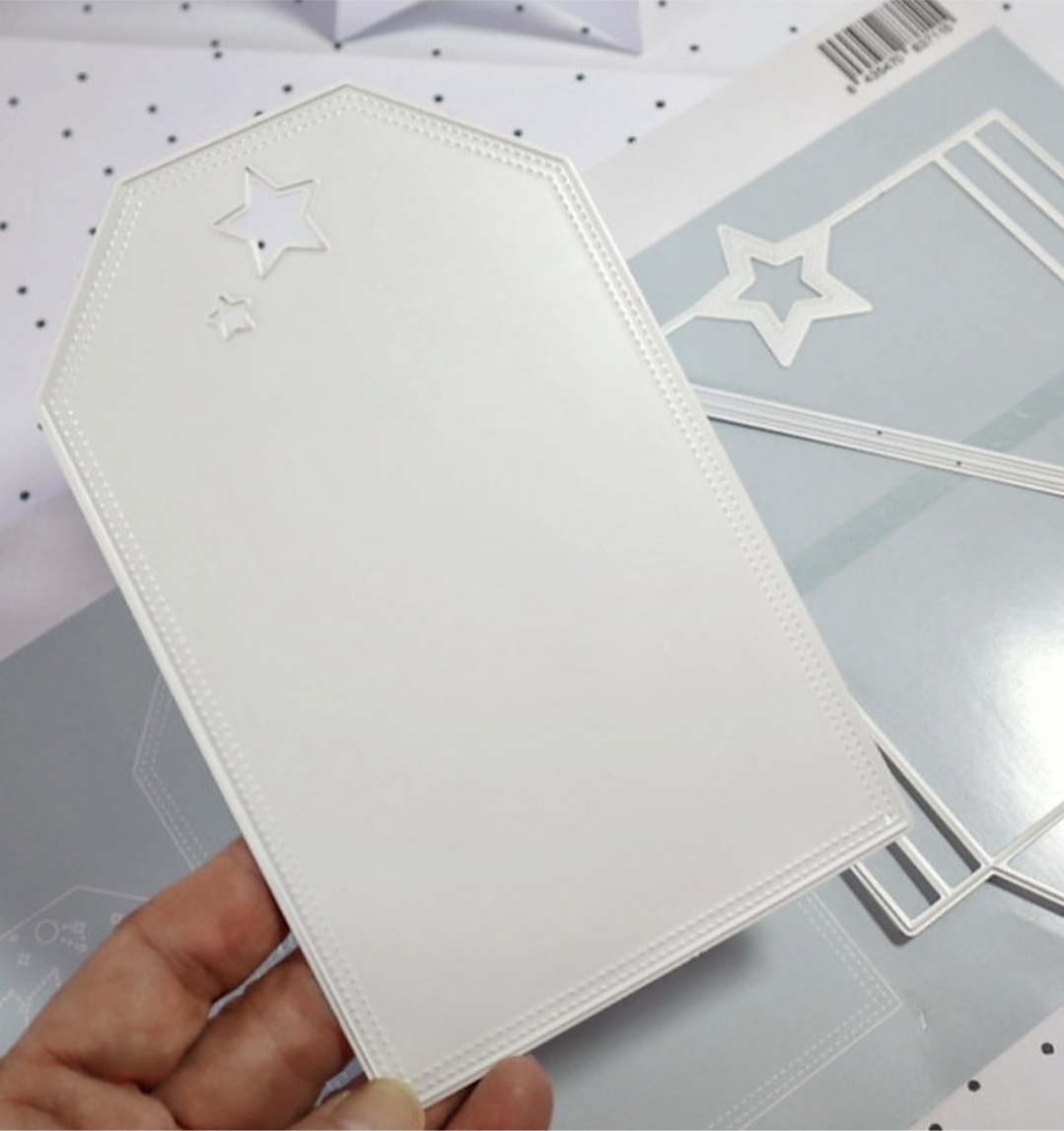Cartão branco em forma de etiqueta com estrelas recortadas e moldes de corte metálicos ao fundo