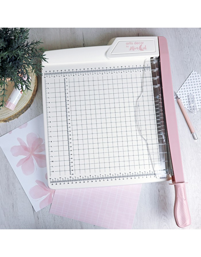 Guilhotina de papel branca e rosa com grelha quadriculada ao lado de folhas e uma planta