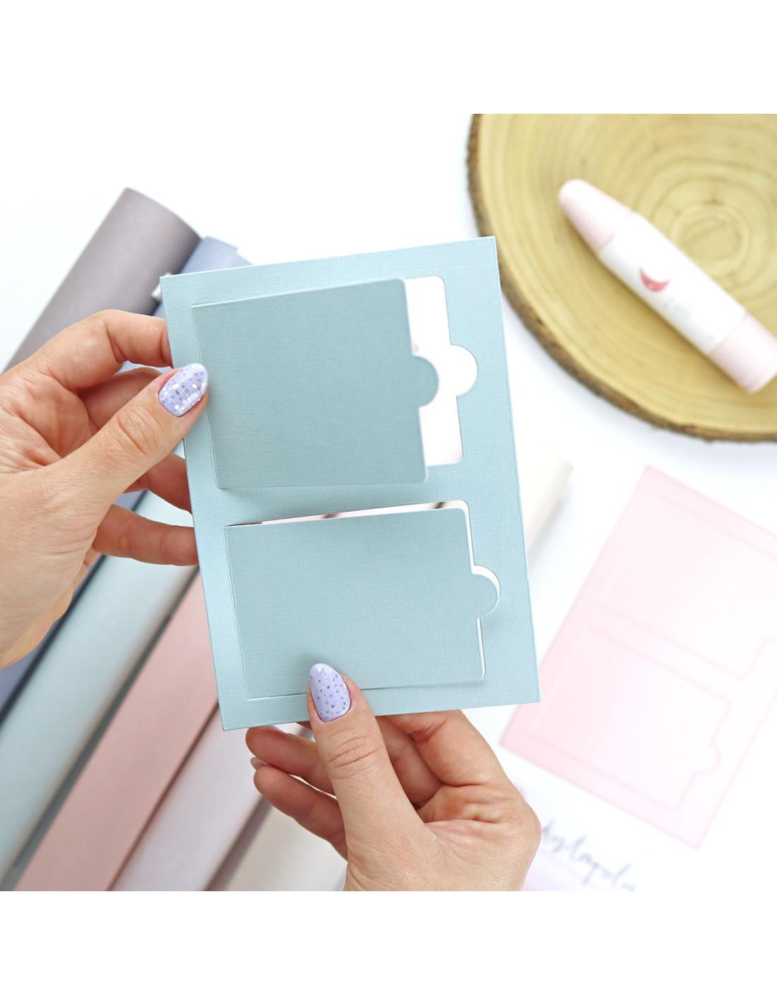 Cartão azul claro com recortes para papel segurado por mãos femininas com unhas pintadas lilás, em mesa com cilindros de papel e batom rosa.