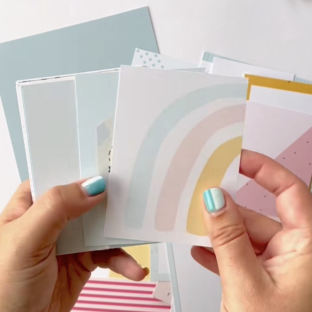 Mãos seguram cartões coloridos com padrões suaves e arco-íris pastel.