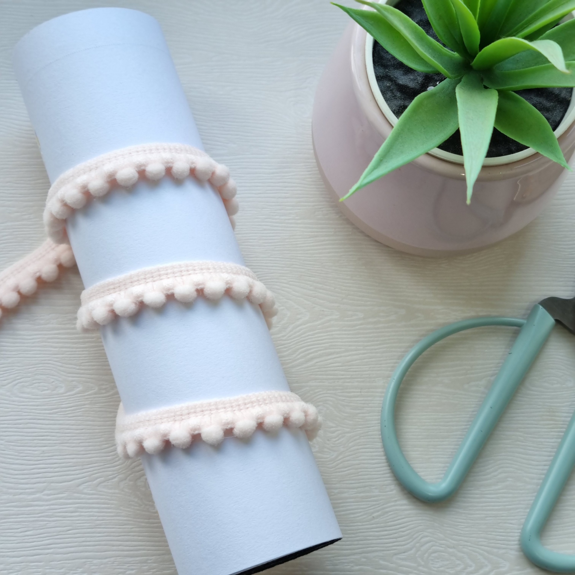 Fita decorativa rosa com pompons junto a planta em vaso e tesoura