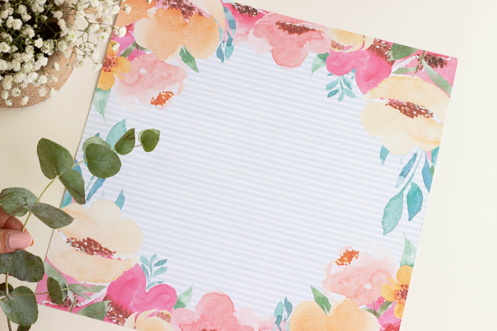 Papel decorativo quadrado com flores coloridas nas bordas e riscas no centro numa mesa clara com planta ao lado