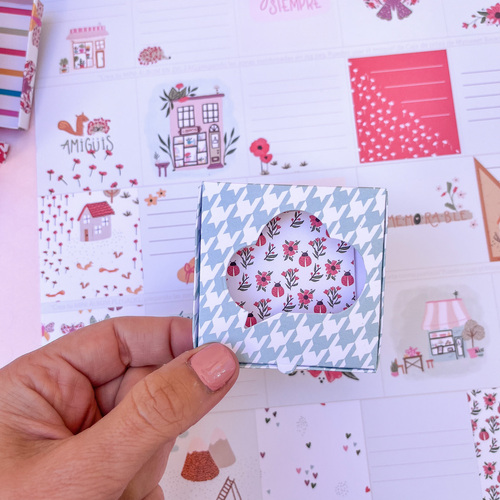 Caixa com padrão pied de poule azul e branco e janela em forma de nuvem com padrão floral vermelho e verde, mão segurando, fundo com cartões decorativos