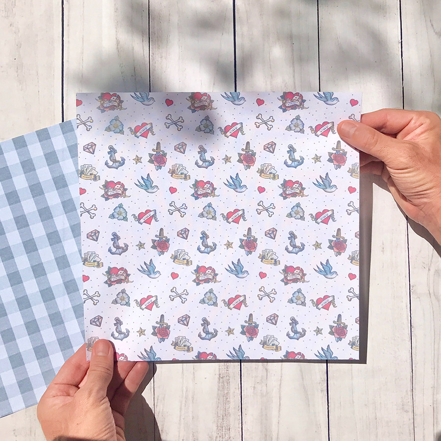 Folha de papel com padrão de tatuagens tradicionais coloridas e folha com padrão de quadrícula azul e branca seguradas sobre mesa de madeira clara