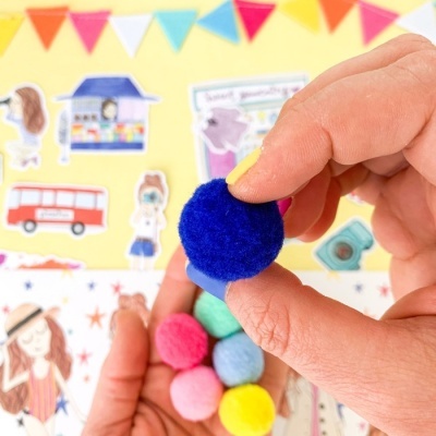 Mãos segurando pompons feltro coloridos com fundo ilustrado amarelo.