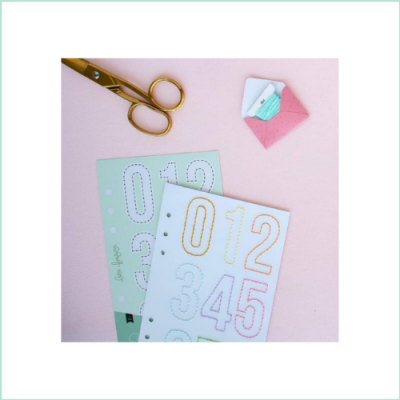 Stencil de números, cartões coloridos, tesoura dourada e pequeno envelope rosa em fundo rosa.
