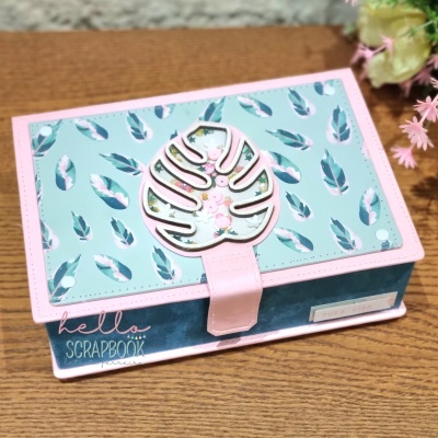 Caixa decorativa azul com padrão de penas e folha rosa em tampa, sobre mesa de madeira