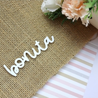 Palavra decorativa 'bonita' em madeira branca sobre juta com flores ao lado