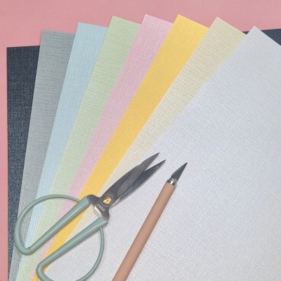 folhas de papel texturizado coloridas, tesoura verde e caneta estilográfica sobre fundo rosa