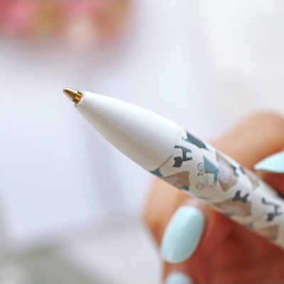 Caneta esferográfica branca decorada com padrão de cães e ponteira dourada, segurada por mão com unhas azuis.