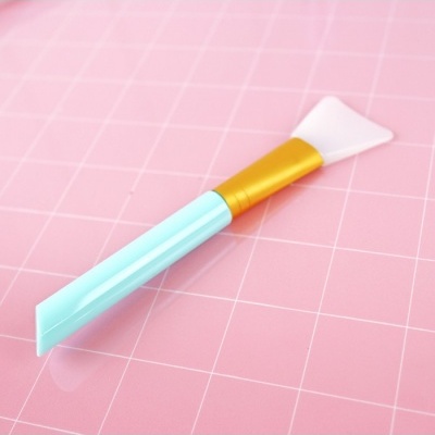 Espátula de silicone azul claro, dourada e branca sobre fundo rosa quadriculado