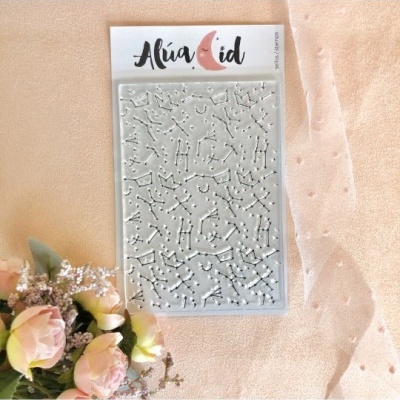 Stencil de constelações em relevo da marca Alua Cid sobre tecido rosa claro próximo a flores.