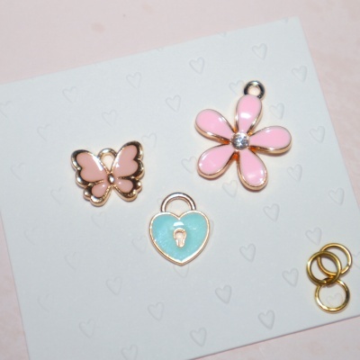 Três pendentes esmaltados coloridos e três argolas douradas, sobre papel com corações em relevo.