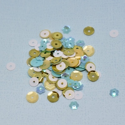 Lantejoulas douradas, brancas e azuis claras em formas redondas e florais sobre superfície azul clara