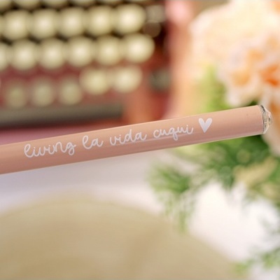 Lápis de cor pêssego com texto 'living la vida cuqui' e coração, fundo desfocado com teclado e flores.