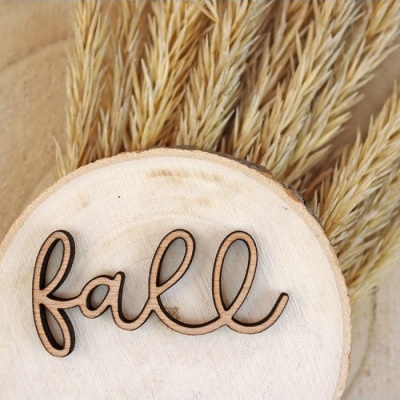 Placa redonda de madeira clara com palavra 'fall' em madeira escura sobre espigas secas