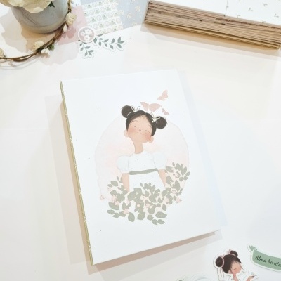 Conjunto de papelaria infantil com caderno, autocolantes e flor numa mesa branca.