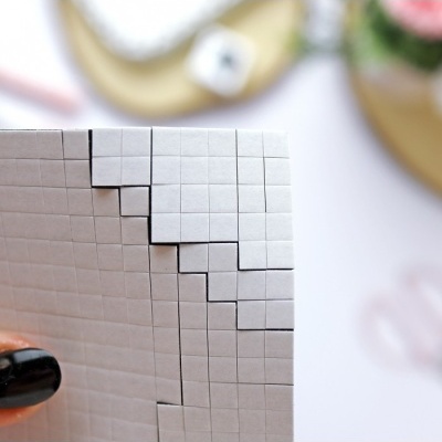 Folha branca quadriculada com recortes em degrau, mão com unhas pretas a segurá-la, fundo desfocado com objetos.