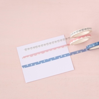 Três rolos de fita adesiva decorativa com corações e um cartão branco sobre superfície rosa