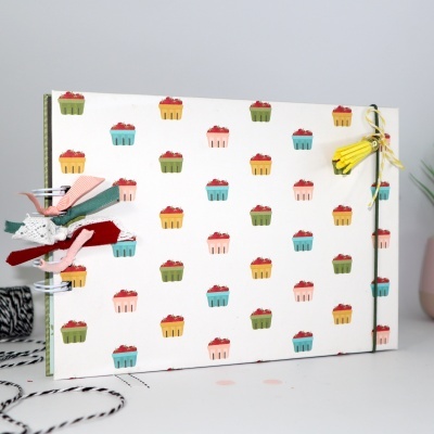 Caderno com capa branca decorada com pequenos cupcakes coloridos e fitas coloridas na encadernação