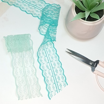 Fitas de renda azul claro e turquesa, tesoura rosa e planta em vaso sobre fundo branco