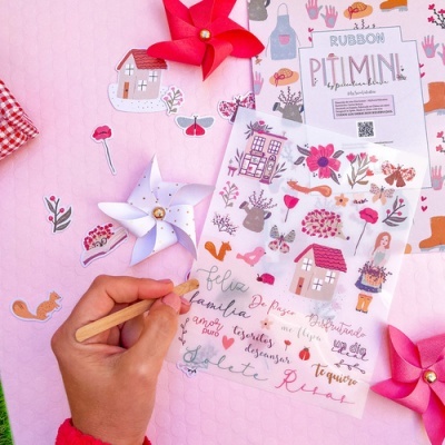 Folha de adesivos decorativos com desenhos coloridos e texto em português sobre mesa rosa com cataventos de papel