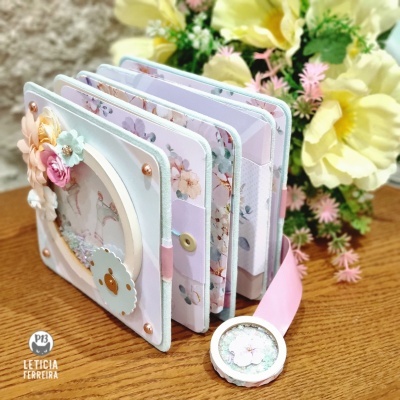 Mini álbum decorado com flores artificiais e papel colorido, com botão e fita rosa, sobre mesa de madeira