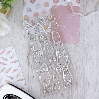 Conjunto de carimbos transparentes de coelhos e itens, sobre mesa de madeira clara com papéis decorativos de fundo.