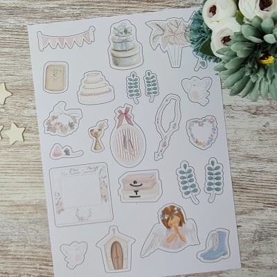 Folha de autocolantes decorativos variados sobre mesa de madeira clara com arranjo floral ao lado.