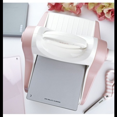 Máquina de corte magnético branca e rosa com unha placa metálica cinza e texto, tesoura rosa listrada e acessórios sobre fundo branco