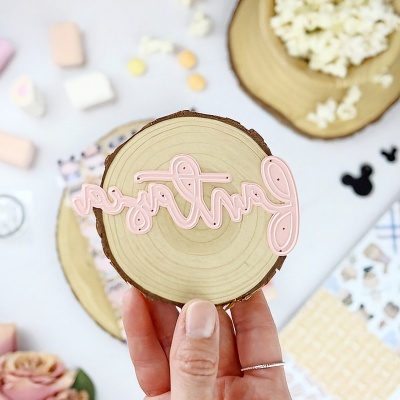 Disco de madeira com palavra rosa 'partners' em relevo, segurado por mão, fundo desfocado com marshmallows e pipoca.