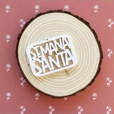 Enfeite branco com 'SEMANA SANTA' sobre uma placa redonda de madeira clara, fundo rosa com flores brancas