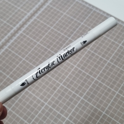 Marcador acrílico branco com tampa branca e texto impresso, sobre superfície quadriculada cinza