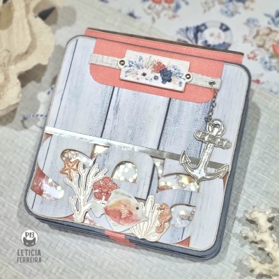 Estojo quadrado com padrão de tábuas brancas e detalhes em coral, com enfeites metálicos de anzol e corais.