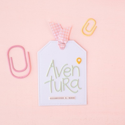 Etiqueta branca em formato de tag com laço xadrez rosa e branco e texto colorido rodeada por clips coloridos sobre fundo rosa claro.