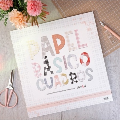 Bloco de papel para scrapbooking com letras coloridas e quadriculadas, tesoura e flores ao fundo.