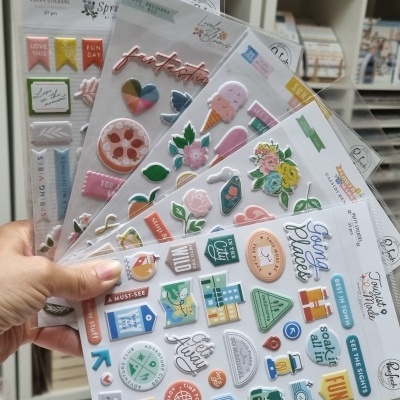 Folhas de autocolantes 3D coloridos com padrões de flores, comida e textos, seguradas por uma mão num ambiente com prateleiras no fundo.