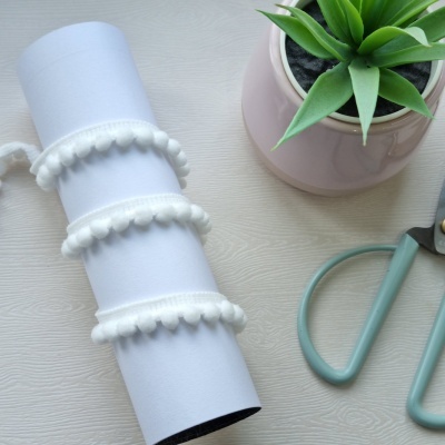 Fita branca com pompons ao lado de planta artificial em vaso rosa e tesoura verde