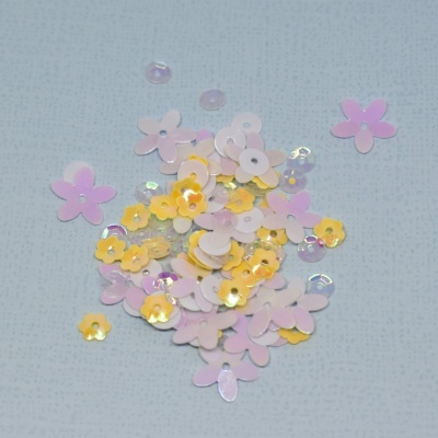 Lantejoulas em forma de flor amarelas, rosa e transparentes sobre fundo cinzento claro