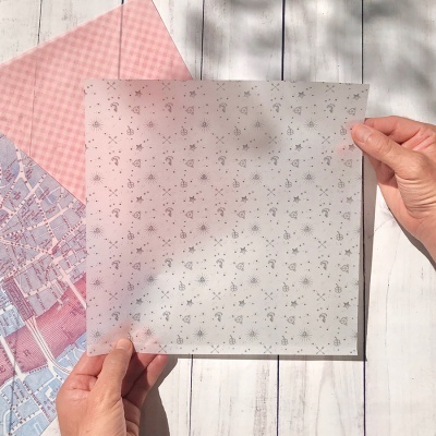 Folha translúcida com padrão floral cinzento sendo segurada por mãos sobre superfície branca