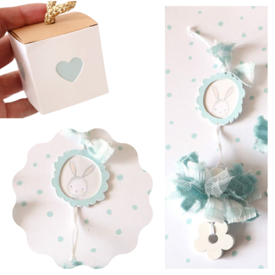Caixa branca com decoração de coração e ornamento pendurado com coelho e fita azul numa superfície com bolinhas azuis.