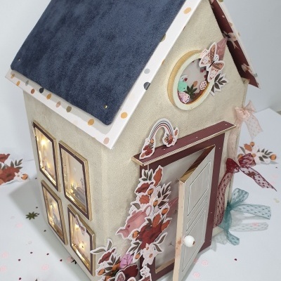 Casa decorativa em tecido cinzento claro com telhado azul e janelas iluminadas