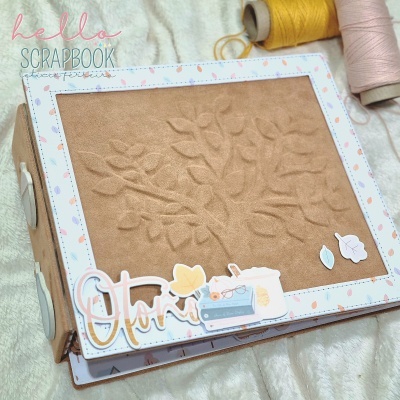 Álbum scrapbook com capa texturizada e decoração em papel, cones de fios ao fundo