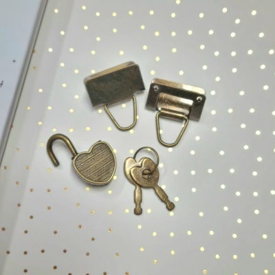 Conjunto de cadeado, chaves e suportes metálicos em bronze sobre fundo branco com pontos dourados