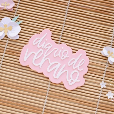 Etiqueta rosa claro com texto 'diário de verão' sobre esteira de madeira clara com flores de papel coloridas